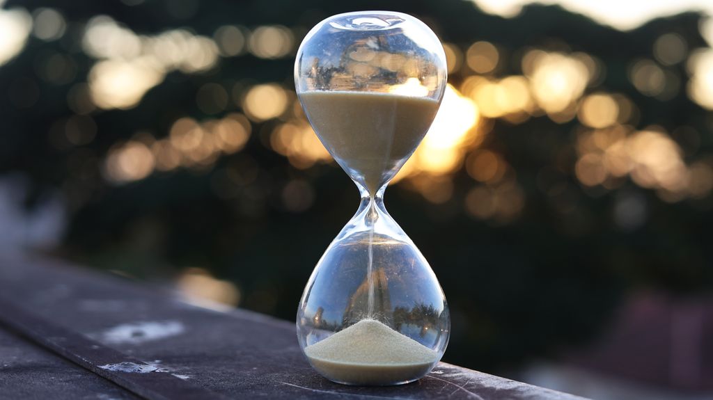 An hourglass with sand on a ledge set against a sunset background, symbolizing time passage.