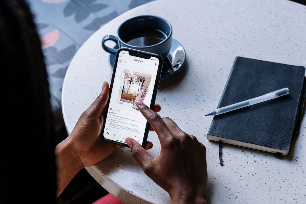Hands holding smartphone at a cafe table with coffee and a notebook, focusing on social media browsing.